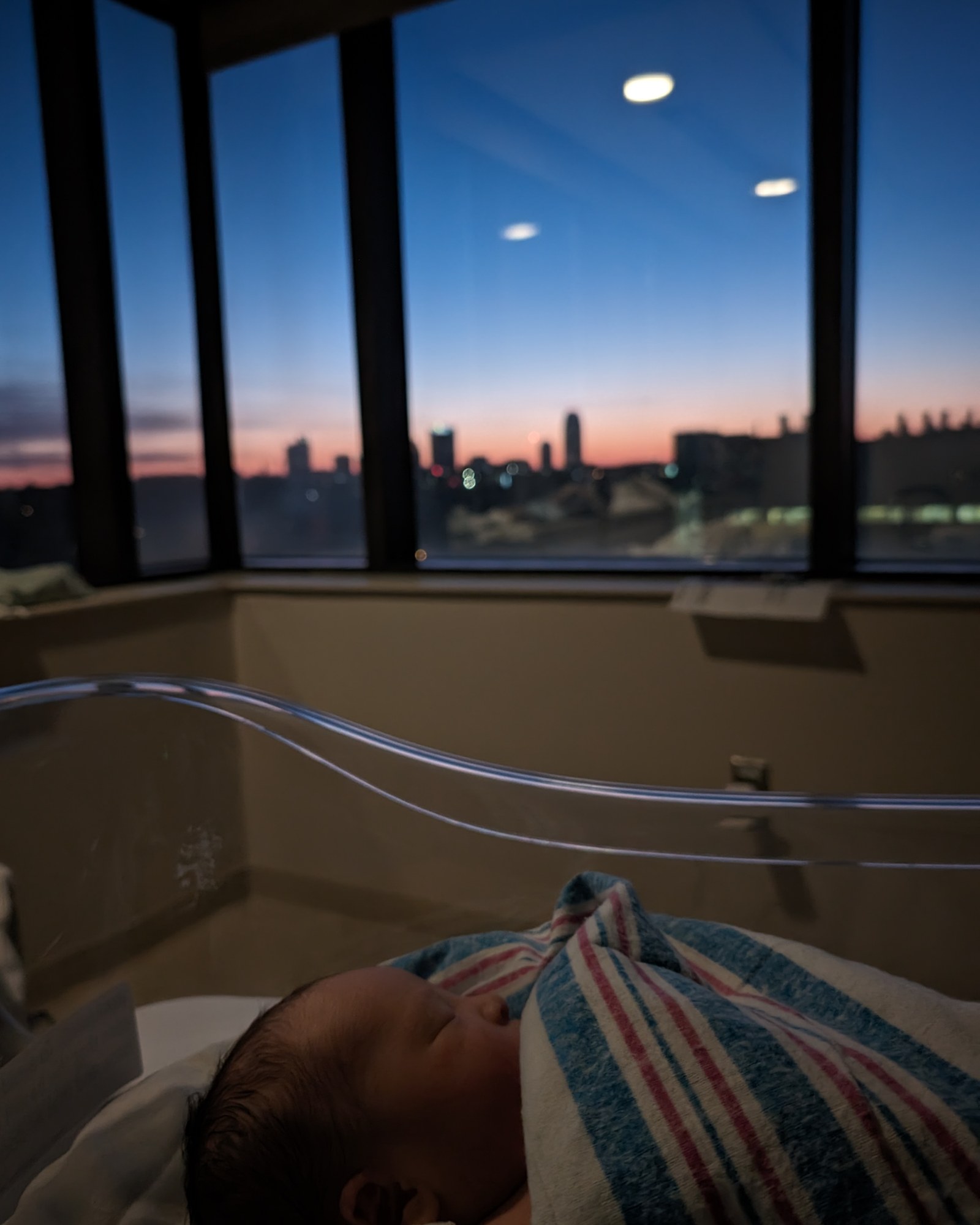 A newborn asleep in the maternity ward, the Winston-Salem skyline lit at dawn through the window